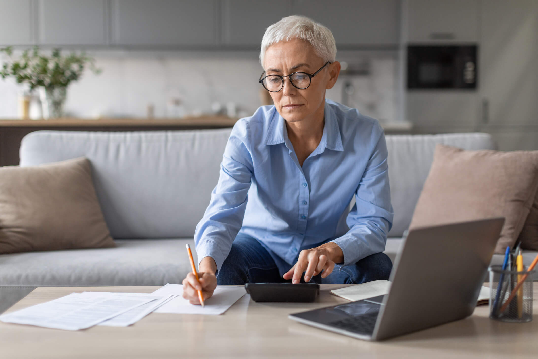 Mature lady entrepreneur in eyewear working with laptop at home and taking notes, businesswoman sitting on sofa at table writing down information, works remotely. Freelance and internet